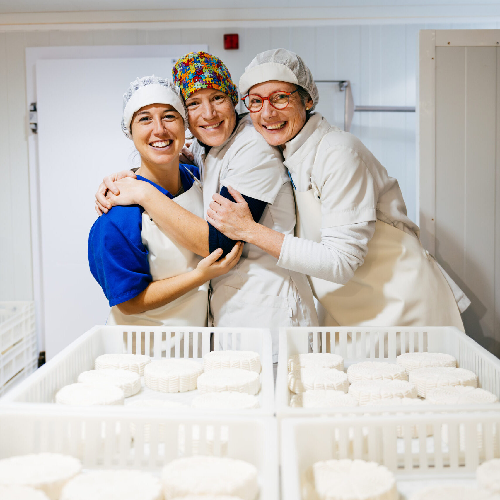 Portrait of smiling factory workers in uniforms hugging each other near white containers with ricotta cheese